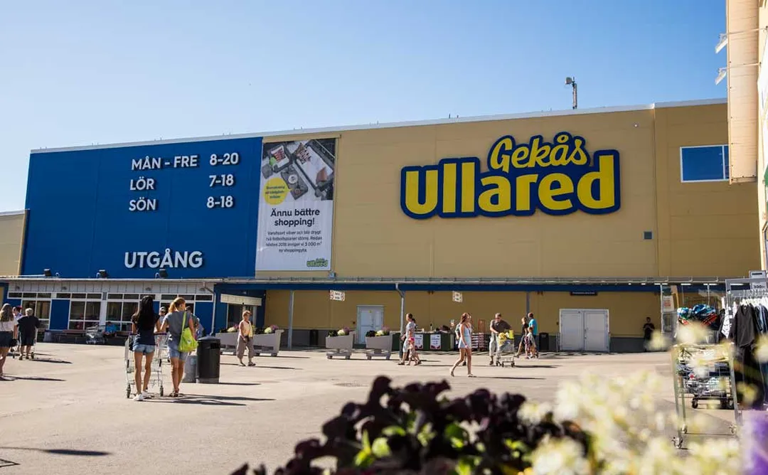 Röd tegelbyggnad med vita fönster bakom urban plats med trappor och moderna silverfärgade skulpturer, röd bil och parkerade cyklar.